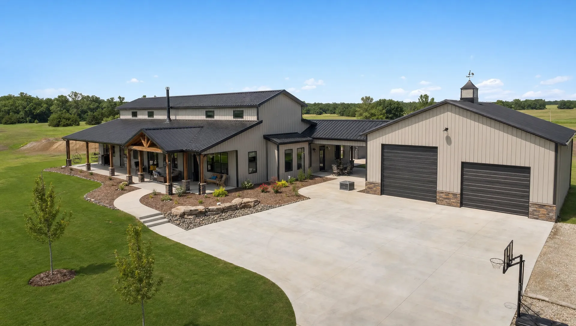 A custom barndominium with an attached covered patio and a nearby metal shop building on a spacious Oklahoma property, showing coordinated concrete flatwork, rooflines, large doors, and outdoor living space from a slightly elevated angle.