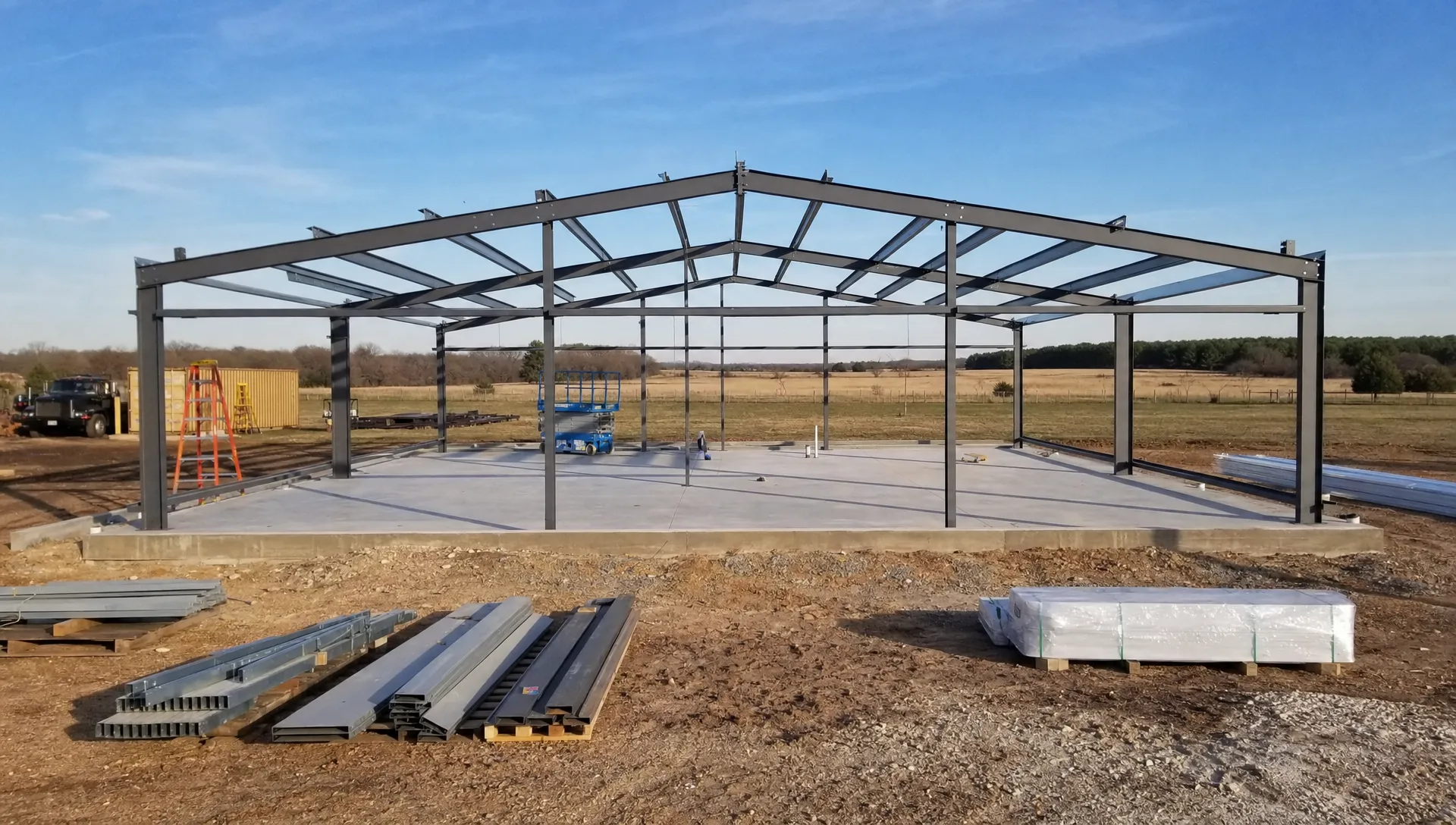 A custom metal building project under construction on rural Oklahoma land, with a completed foundation, framed structure, organized materials, and open acreage in the background.