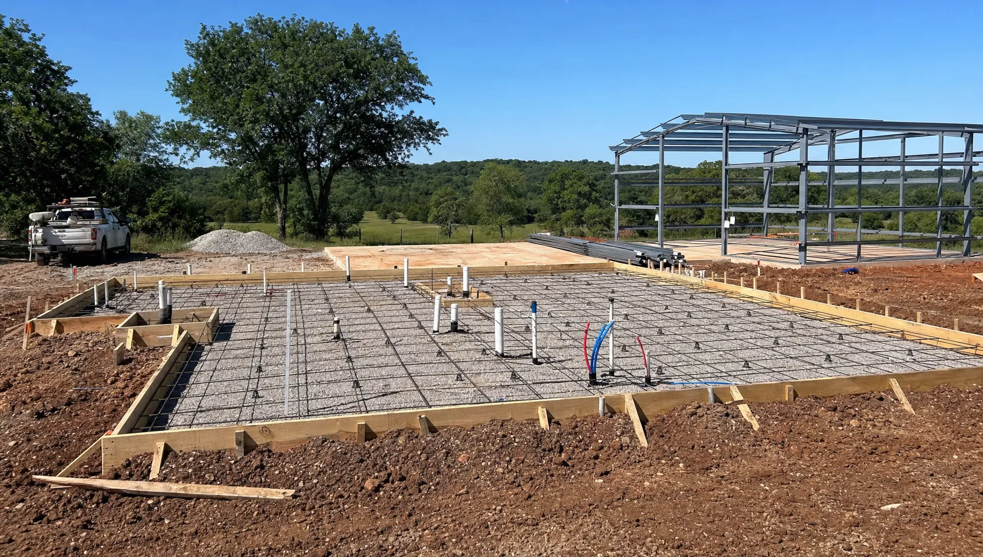 A prepared foundation site in northeast Oklahoma with forms, compacted base, rebar on chairs, plumbing rough-ins, and a metal building frame staged nearby on rural land.