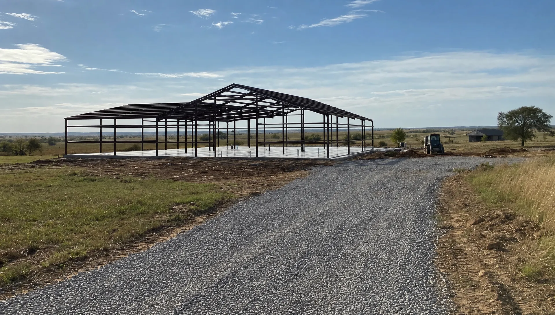 A rural Oklahoma barndominium site with a metal building frame on a concrete slab, a gravel access drive, and open prairie around the property under a wide sky.