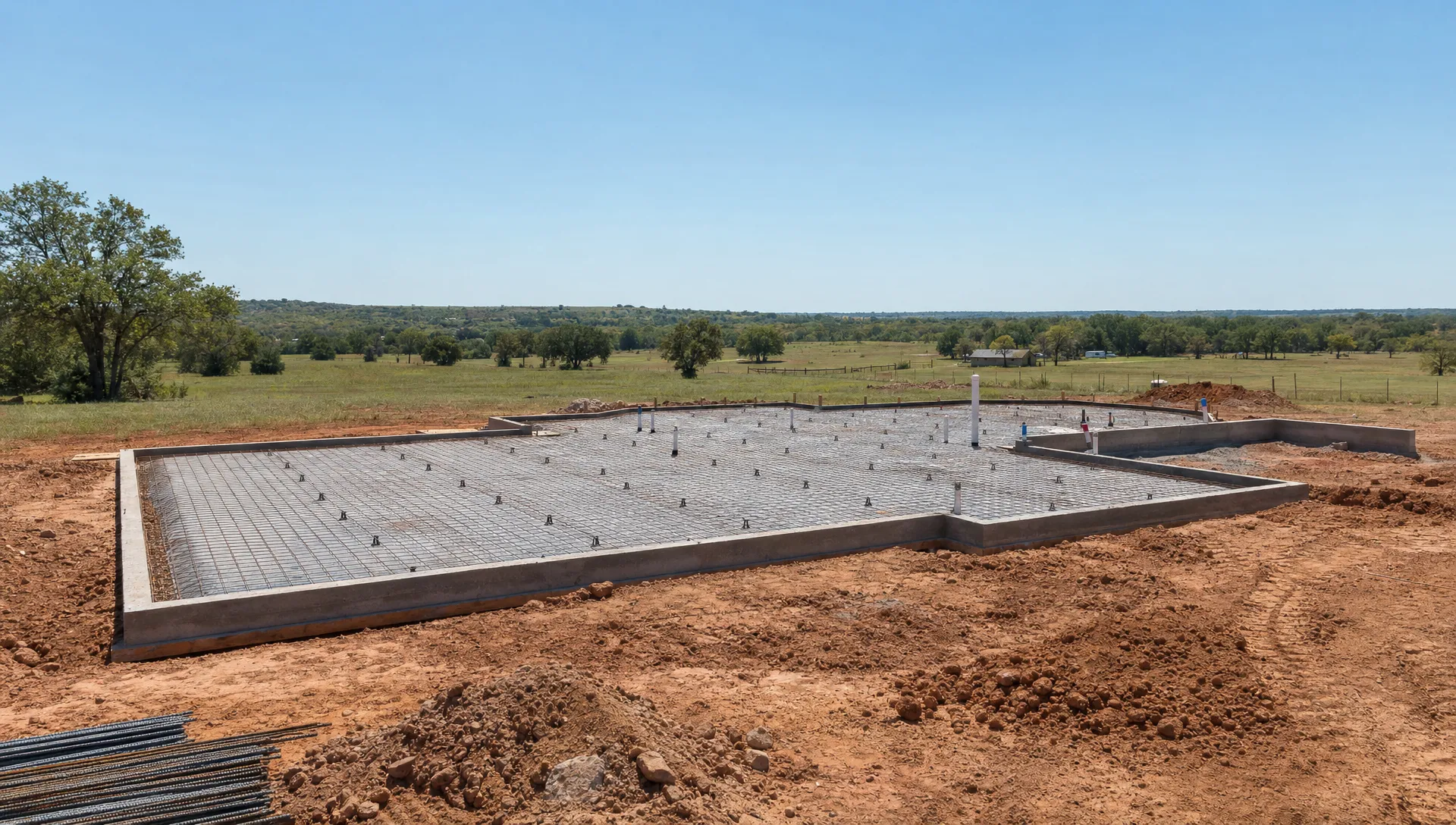 An Oklahoma acreage construction site with a prepared building pad, reinforced concrete foundation work in progress, and open rural land surrounding the future barndominium home.